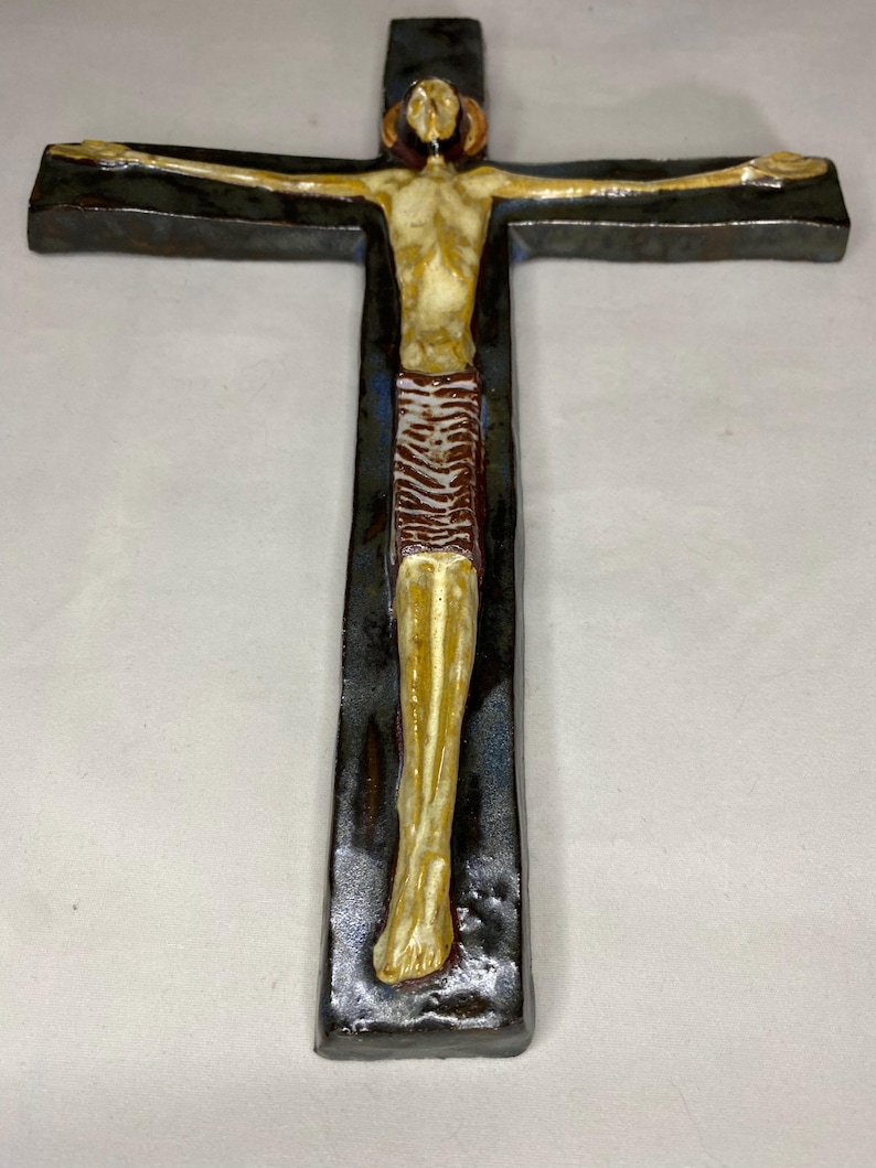 Może przedstawiać: Ceramiczny krzyż ścienny z ciemnoniebieską glazurą i beżową figurą Jezusa Chrystusa. Figura jest przedstawiona w brązowym przepasaniu i z wyciągniętymi ramionami.