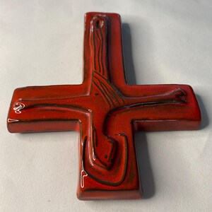 May include: A red ceramic cross with a glossy finish. The cross features a stylized design with dark brown lines and a hole at the top for hanging. The cross is a religious symbol.