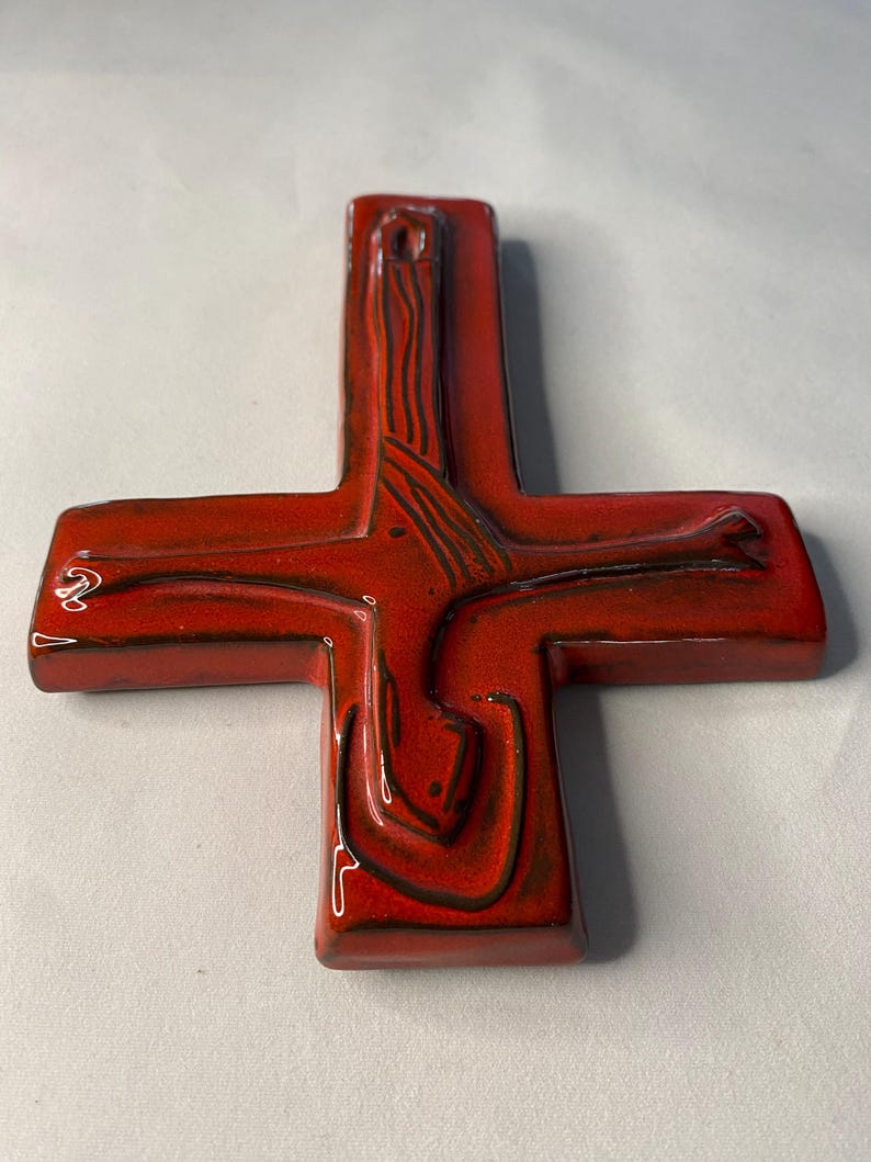 May include: A red ceramic cross with a glossy finish. The cross features a stylized design with dark brown lines and a hole at the top for hanging. The cross is a religious symbol.