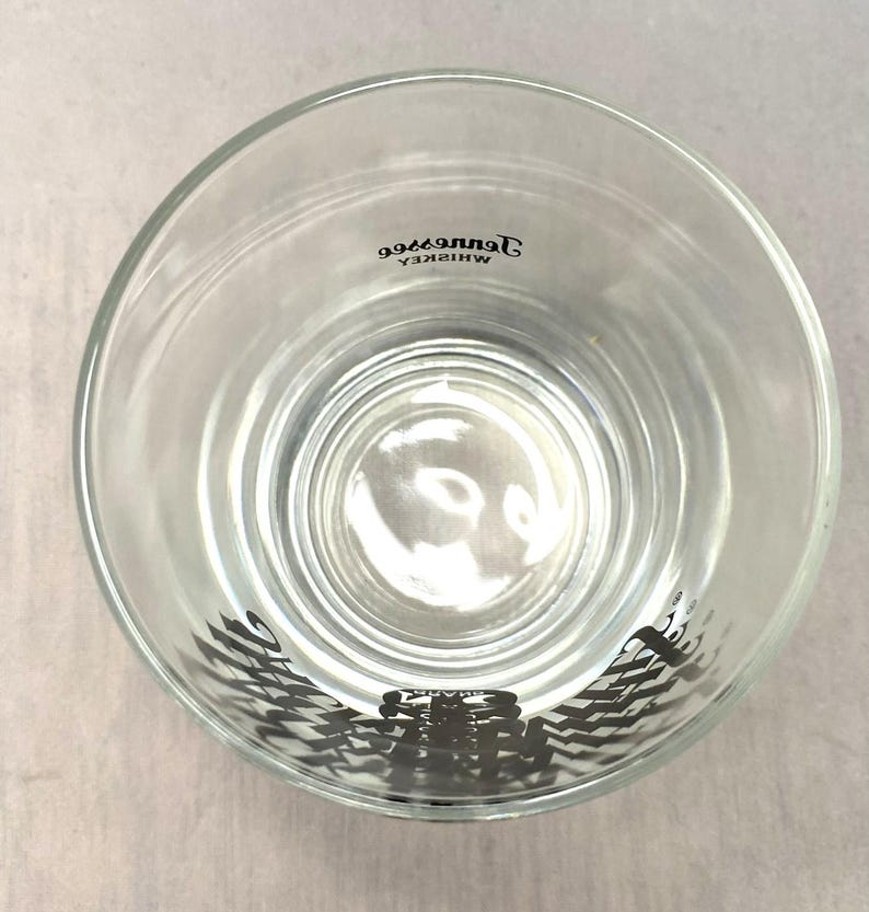 May include: Clear, round whiskey glass with a thick base. The glass has the words "Tennessee Whiskey" printed in black, and a black graphic design at the bottom. The glass is empty.