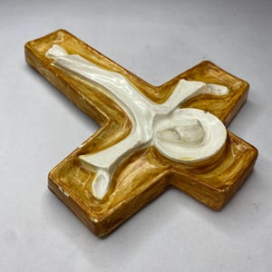 May include: A ceramic cross with a figure of Jesus. The cross is a warm, golden-brown color with a white figure in the center. The figure is in a reclining position, with arms outstretched. The cross is a religious item.