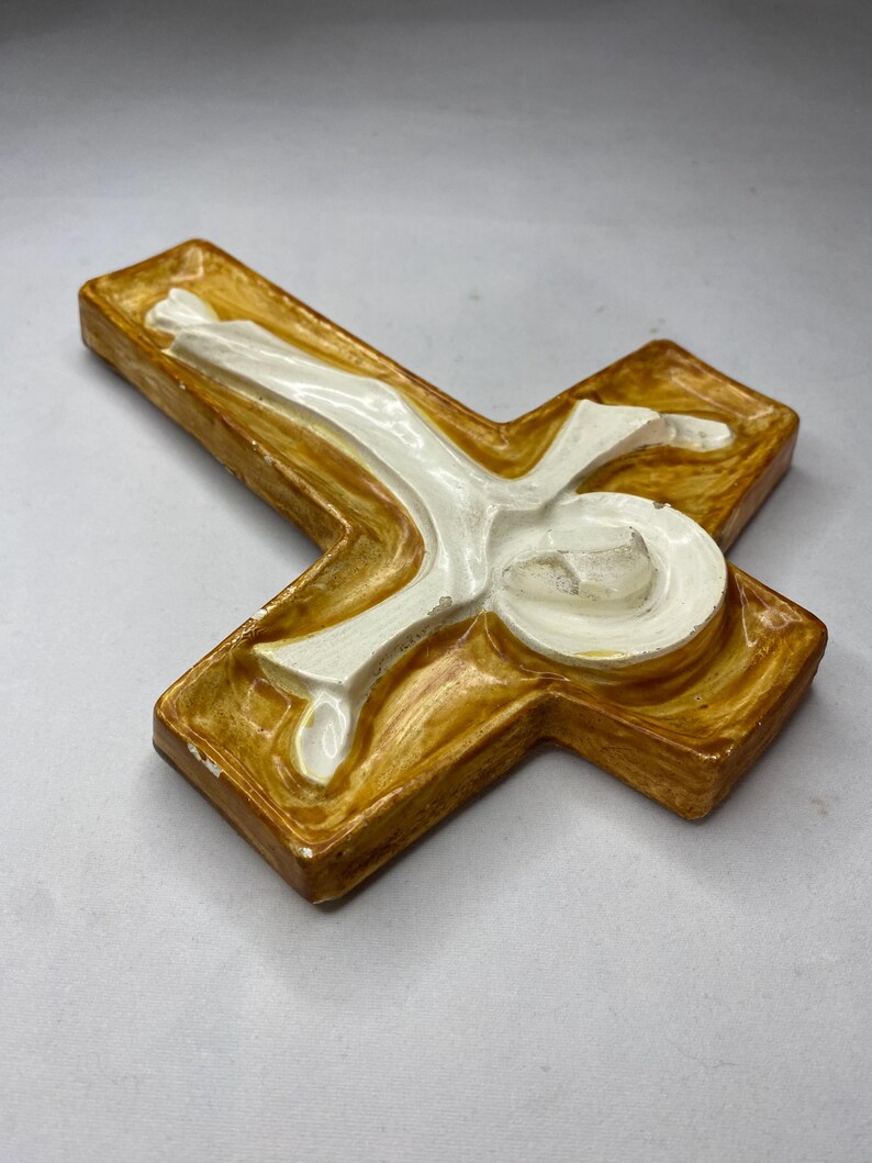May include: A ceramic cross with a figure of Jesus. The cross is a warm, golden-brown color with a white figure in the center. The figure is in a reclining position, with arms outstretched. The cross is a religious item.