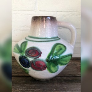 Peut inclure: Un vase en céramique blanche avec un glaçage brun sur le dessus et un motif floral vert et rouge. Le vase a une anse et un corps arrondi.