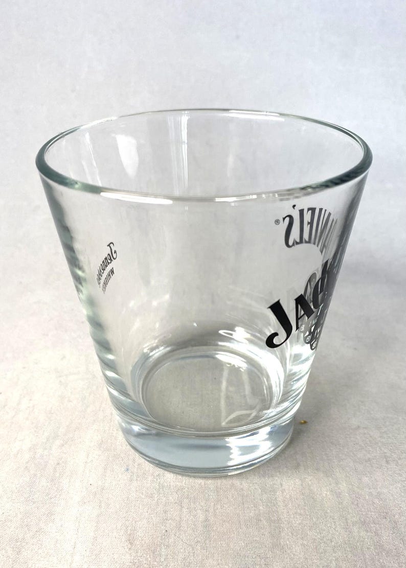 May include: A clear shot glass with a tapered design, featuring the "JACK DANIEL'S" logo in black. The glass has a sturdy base and a slightly flared rim. The logo is printed on the side. Ideal for serving spirits, this glass is a classic.