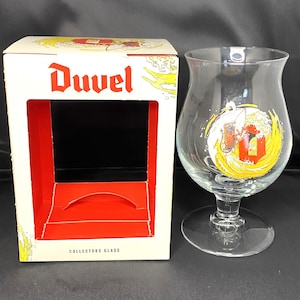 May include: A white box with red trim and the word "Duvel" printed in red. The box is labeled "Collectors Glass". A clear glass with a yellow and red design is next to the box.