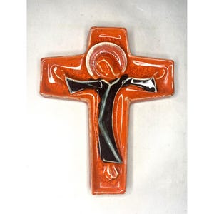May include: Orange ceramic cross with a black and white figure of a person with outstretched arms. The figure is surrounded by a red halo.