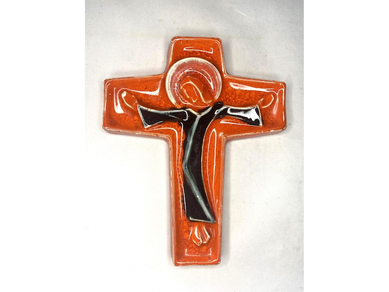 May include: Orange ceramic cross with a black and white figure of a person with outstretched arms. The figure is surrounded by a red halo.