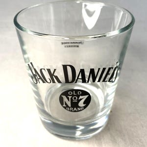 May include: A clear glass shot glass featuring the "Jack Daniel's" logo in black. The logo includes the words "Old No. 7 Brand" in a circular design. The glass has a cylindrical shape with a slightly flared top.