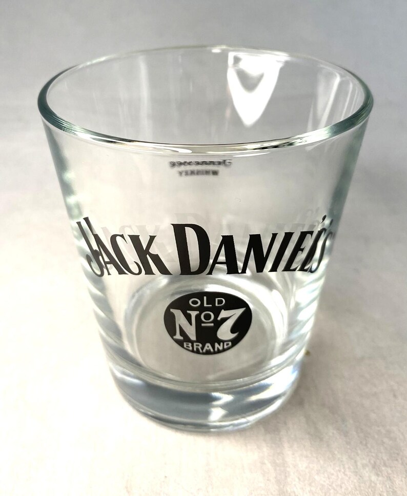 May include: A clear glass shot glass featuring the "Jack Daniel's" logo in black. The logo includes the words "Old No. 7 Brand" in a circular design. The glass has a cylindrical shape with a slightly flared top.