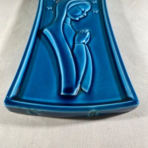 May include: A blue ceramic wall plaque with a raised image of a woman in a robe with her hands clasped in prayer. The background is decorated with white stars.