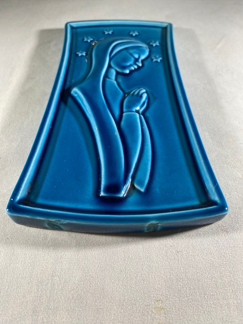 May include: A blue ceramic wall plaque with a raised image of a woman in a robe with her hands clasped in prayer. The background is decorated with white stars.