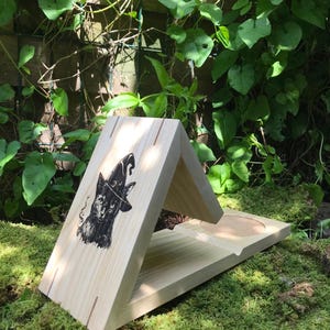 Peut inclure: Un support de téléphone en bois clair avec un motif de chat noir portant un chapeau de sorcière. Le support est triangulaire avec une base plate et une découpe circulaire. Il est posé sur de la mousse verte.