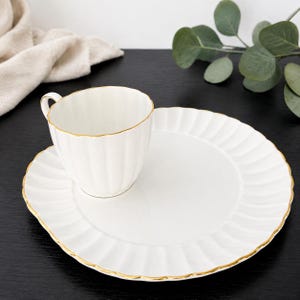 May include: A white ceramic teacup and matching saucer set featuring a delicate fluted design. Both the cup and the saucer are finished with a thin, elegant gold trim along the scalloped edges, providing a clean and refined appearance for serving.