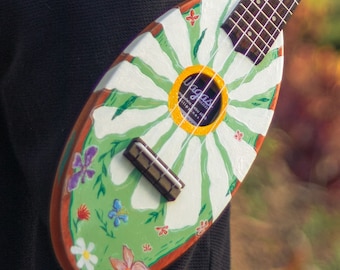 Könnte beinhalten: Bemalte Ukulele mit Gänseblümchen-Design. Der ovale Korpus hat einen grünen Hintergrund und weiße Gänseblümchenblätter. Das Schallloch trägt den Text "Vagas". Die Ukulele ist mit Blumen und Schmetterlingen verziert.