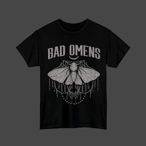 以下が含まれることがあります： ライトグレーのグラフィックプリントが施された黒の半袖Tシャツ。デザインには「BAD OMENS」という文字と、複雑な羽の模様を持つ蛾のイラスト、そしてその下に小さな点が連なる装飾的な垂れ下がった線が描かれています。