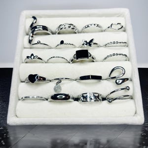 Könnte beinhalten: Eine Sammlung von Silberringen, die in einer weißen Schmuckschatulle ausgestellt sind. Die Ringe weisen verschiedene Designs auf, darunter eine Schlange, Kettenglieder, Herzen und geometrische Formen. Die Ringe sind aus glänzendem Metall gefertigt und ordentlich angeordnet.