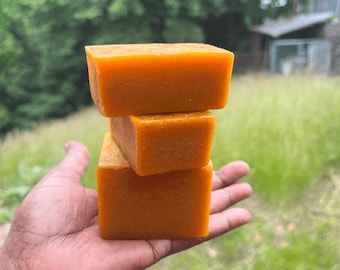May include: Three stacked orange bars of soap. The soap is in the palm of a hand.