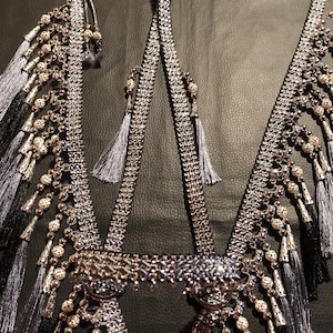 Op de afbeelding: Een sierlijk zilver-grijs decoratief harnas met kwastjes en kralenaccenten. Het harnas heeft ingewikkeld metaalwerk en is versierd met hangende kralen en kwastjes. Het ontwerp omvat een centraal stuk en banden, allemaal in zilver- en grijstinten.