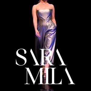 May include: A woman wearing a silver jumpsuit with a strapless neckline and a fitted silhouette. The model is walking on a runway with the text "SARA MILA" in white letters in the foreground.