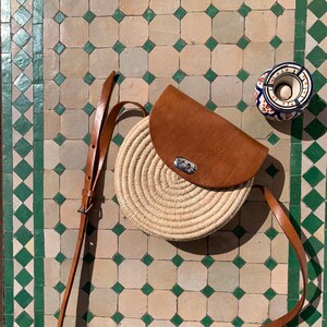 This rounded shoulder bag is hand-woven from natural raffia. It's enriched with high-end vegan leather details and has a pocket for securing small items.