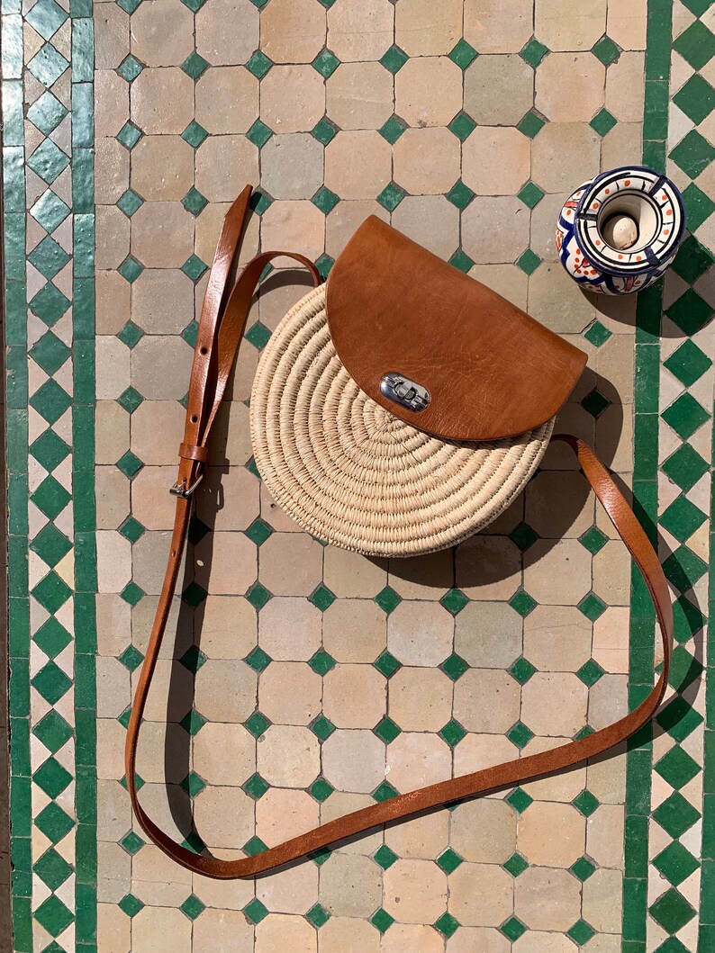 This rounded shoulder bag is hand-woven from natural raffia. It's enriched with high-end vegan leather details and has a pocket for securing small items.