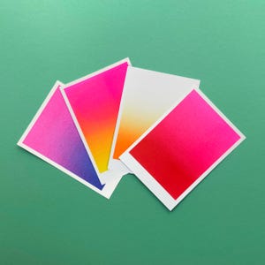 Könnte beinhalten: Eine Anordnung von Karten mit Farbverläufen in Pink, Orange, Gelb und Lila auf einem grünen Hintergrund. Jede Karte hat einen weißen Rand. Die Karten sind fächerförmig angeordnet und zeigen die Farbübergänge.