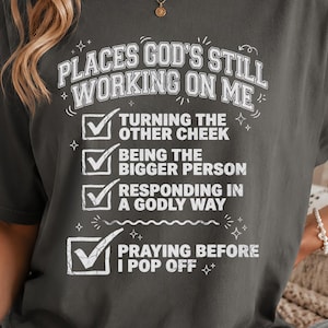 May include: Dark gray t-shirt with the text "PLACES GOD'S STILL WORKING ON ME" and a list of goals. The shirt has a relaxed fit and is made of a soft material. The design includes checkmarks next to each goal.