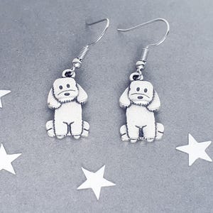 May include: A pair of silver-coloured dog-shaped drop earrings. Each earring features a small, detailed poodle charm suspended from a silver hook. The earrings are displayed on a grey surface with white star-shaped decorations.