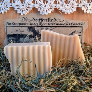 Puede incluir: Dos barras de jabón hecho a mano, una blanca y otra beige, con una textura acanalada, sentadas sobre un lecho de hierba seca. El jabón está etiquetado como "De Seifentiede" con el texto "Ein Sonnenhut-Lundes Wort treibt manches Dante kot"