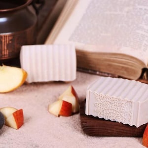 May include: Two bars of white soap, one with a decorative pattern, on a wooden surface with a book and apples.