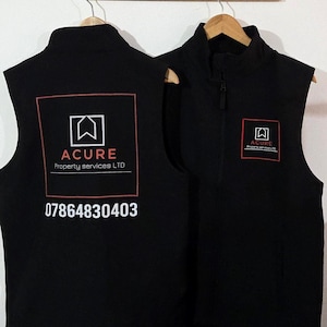 Può includere: Due gilet in pile nero con un logo rosso che dice "ACURE property services LTD" e un numero di telefono. Il gilet a sinistra ha il logo e il numero di telefono stampati sul retro. Il gilet a destra ha il logo stampato sul davanti.