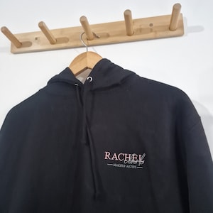 May include: Black hooded sweatshirt with a pink embroidered logo that says "Rachel Clark - Makeup Artist".