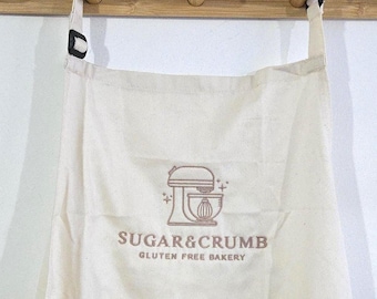 May include: Off-white apron with the text "SUGAR & CRUMB GLUTEN FREE BAKERY" and a graphic of a mixer. The apron is hanging on a wooden rack with three pegs. The apron has black straps and a pocket.