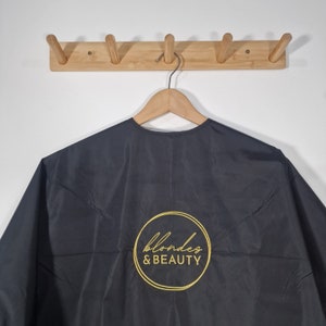 Può includere: Grembiule da taglio nero con un logo circolare dorato che dice "blondes & BEAUTY".