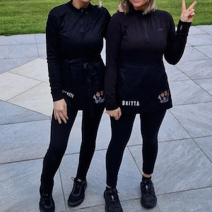 Puede incluir: Dos personas con camisas de cuello alto negras, leggings negros y zapatos negros. Ambas llevan delantales negros con texto blanco que dice "Britta" y "Annette".