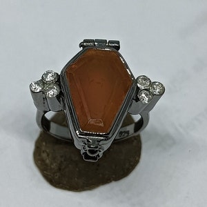 May include: A black ring with a coffin-shaped compartment and a clear orange stone. The ring is decorated with small clear stones.