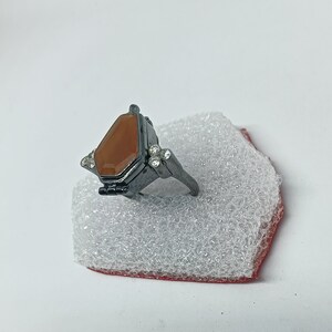May include: A black ring with a brown stone set in a coffin-shaped design. The ring has small clear stones on the sides.