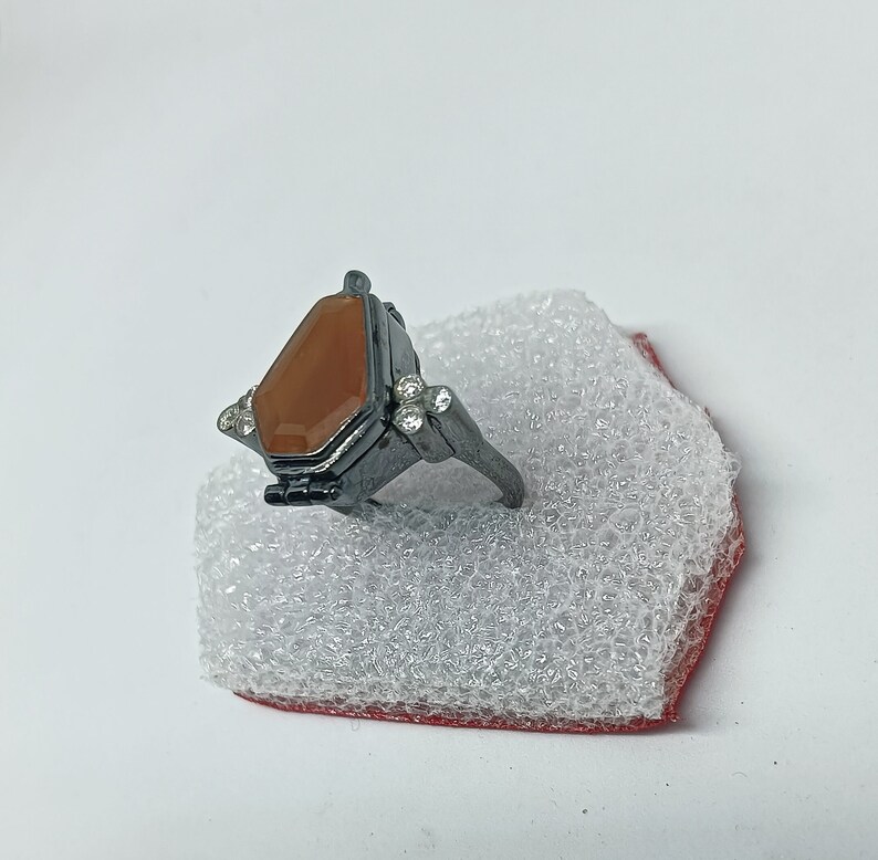 May include: A black ring with a brown stone set in a coffin-shaped design. The ring has small clear stones on the sides.