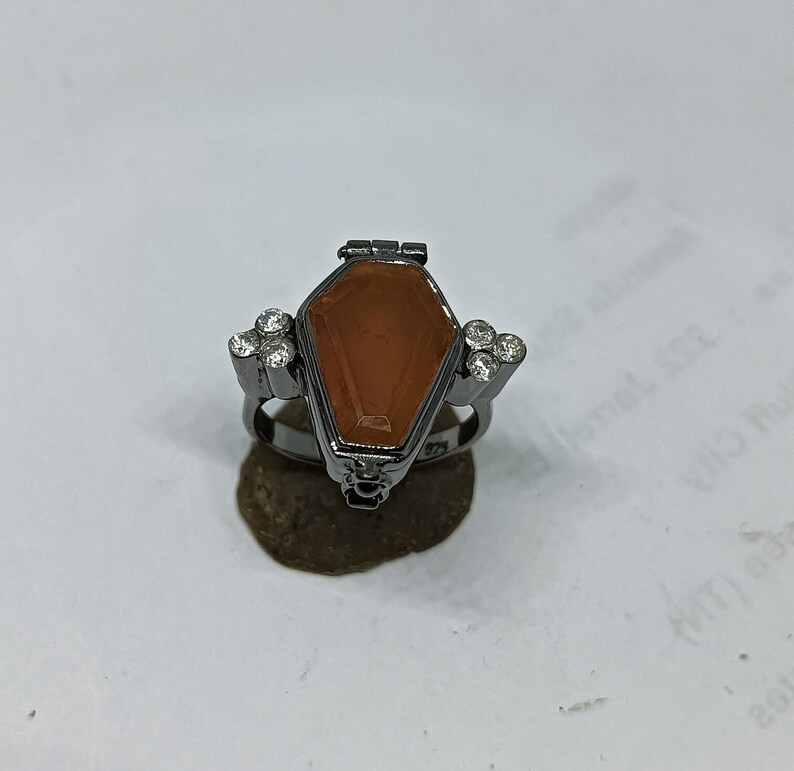May include: A black ring with a coffin-shaped compartment and a clear orange stone. The ring is decorated with small clear stones.