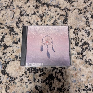May include: A CD case featuring a dreamcatcher design on a pink and purple background. The dreamcatcher is detailed with a web, beads, and feathers. The text "70MM SIX TRACK" and "DOLBY STEREO" is visible on the bottom left.