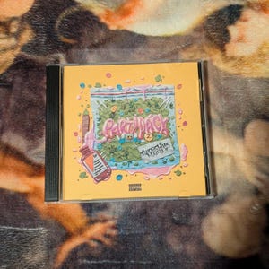 Op de afbeelding: Een cd-hoesje met een gele cover. De illustratie toont de tekst "Party Pack" in roze en blauwe letters, een doorzichtige zak met groene planten, een fles en de tekst "Shoreline Mafia". De hoes zit in een doorzichtige plastic hoes.