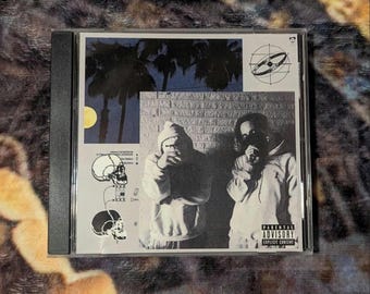 May include: A CD case with a black spine and a white front cover. The cover features a black and white photo of two people wearing hooded sweatshirts. The background is a brick wall with a palm tree and a blue sky. The text "Parental Advisory Explicit Content" is printed in white on a black rectangle at the bottom right corner of the cover.