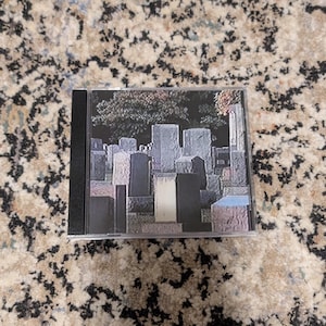May include: A CD case featuring an album cover with a cemetery scene. The image shows various headstones in shades of gray, black, and white, with trees in the background. The CD case is black with a clear plastic front.