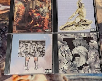 Puede incluir: Cuatro discos compactos con diferentes ilustraciones. El primer disco presenta una pintura de una escena bíblica. El segundo disco presenta una escultura de un dios griego. El tercer disco presenta una pintura de una escena de batalla. El cuarto disco presenta un diseño gráfico de un casco.