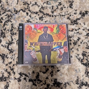 May include: A compact disc case featuring the album cover for "WhiteRapper." The cover art includes a central figure in a black outfit, surrounded by images of other people. The album title is prominently displayed in yellow. The CD case is black.