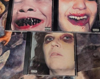 Può includere: Tre dischi compatti con immagini grafiche di volti di persone. Il primo disco presenta un primo piano di un viso con un ampio sorriso e denti neri. Il secondo disco presenta un primo piano di un viso con un ampio sorriso e sangue intorno alla bocca. Il terzo disco presenta un primo piano di un viso con un occhio nero e un'espressione seria. Tutti e tre i dischi hanno un adesivo di avvertenza per i genitori.