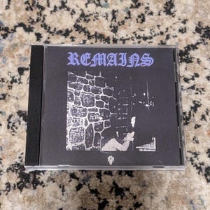 May include: A CD case with the word "REMAINS" in a gothic font. The cover art is a black and white illustration of a stone wall and a figure. The CD case is in a clear plastic sleeve.