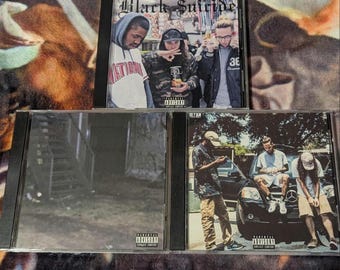 Può includere: Tre dischi compatti con custodie nere e etichette di avvertenza per i genitori. La copertina anteriore del CD superiore presenta un gruppo di tre uomini con il testo "Black Suicide" in bianco e nero. La copertina anteriore del CD centrale è un'immagine sfocata di un edificio con una scala. La copertina anteriore del CD inferiore presenta tre uomini in piedi davanti a un'auto nera con il testo "Parental Advisory" in bianco e nero.