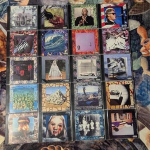 Op de afbeelding: Een verzameling van zestien cd's, elk in een jewelcase, met albumhoezen met diverse afbeeldingen. De kunstwerken omvatten portretten, landschappen en abstracte ontwerpen. Sommige cases tonen een waarschuwing voor ouders.