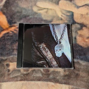 May include: A CD case featuring a close-up image of a person wearing a diamond-encrusted skull pendant necklace. The person is wearing a jacket with a camouflage pattern.
